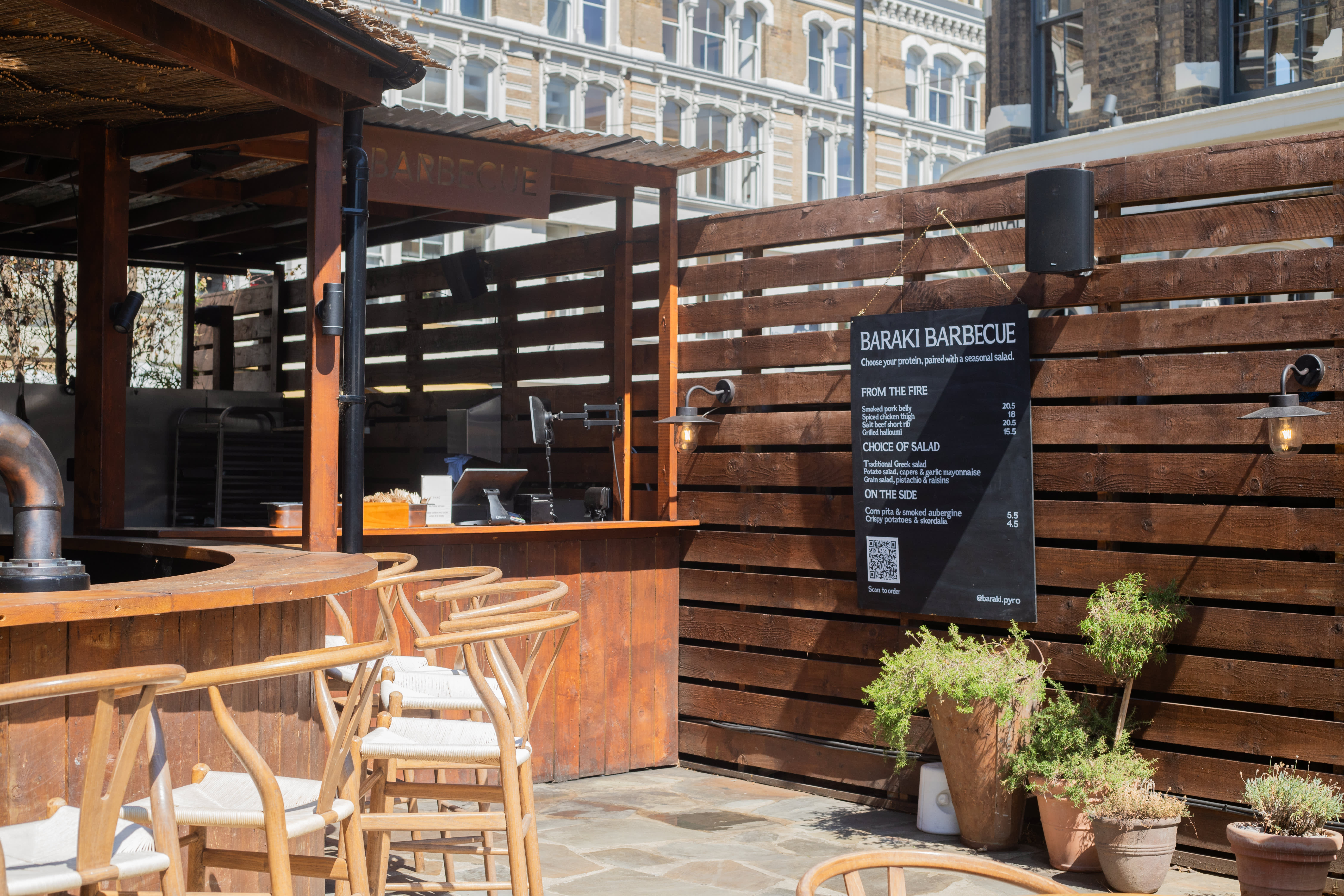 A hand-painted menu board on a timber fence beside outdoor bar seating.